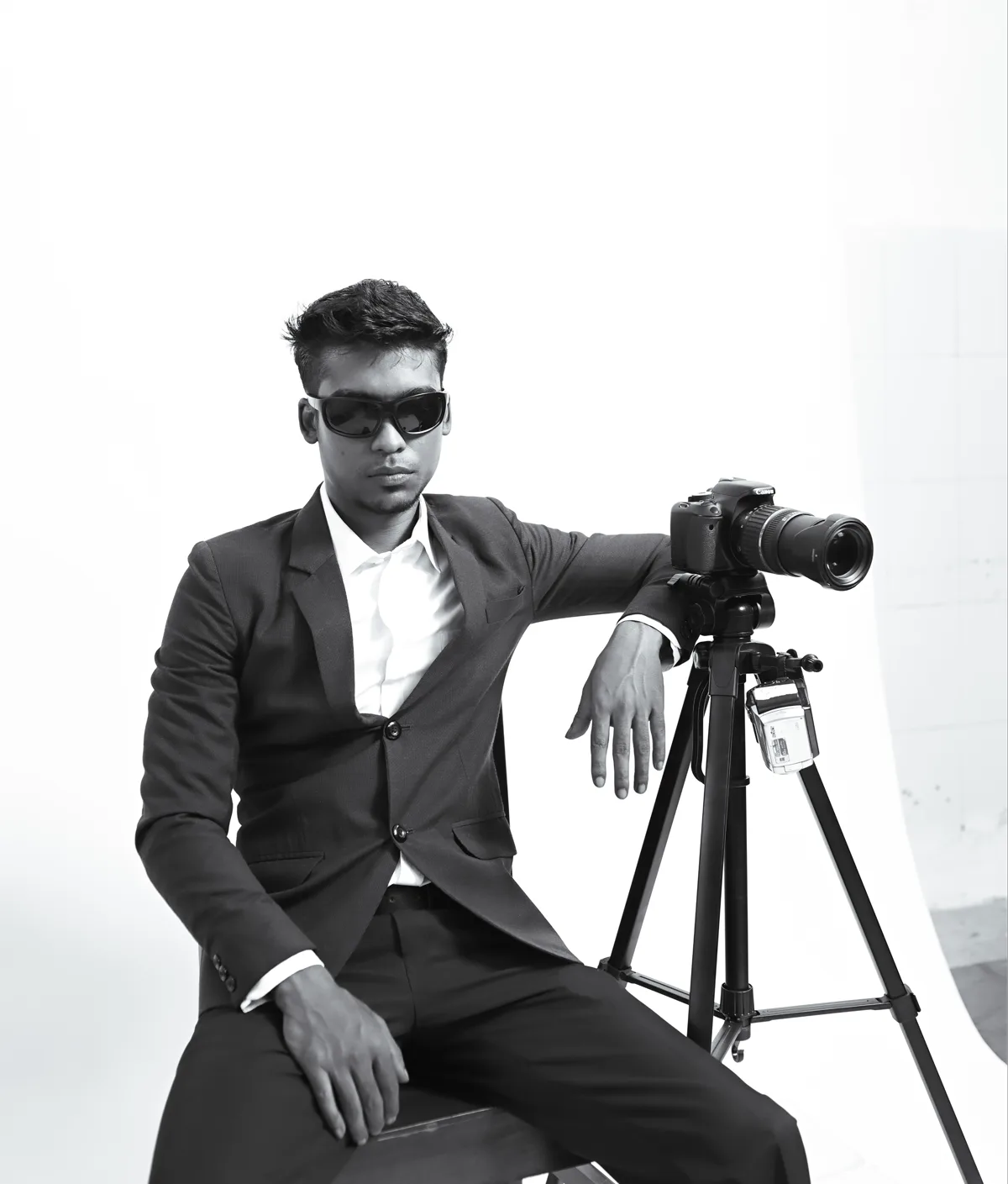 Amrit Xess seated beside a camera on a tripod, photographed in black and white