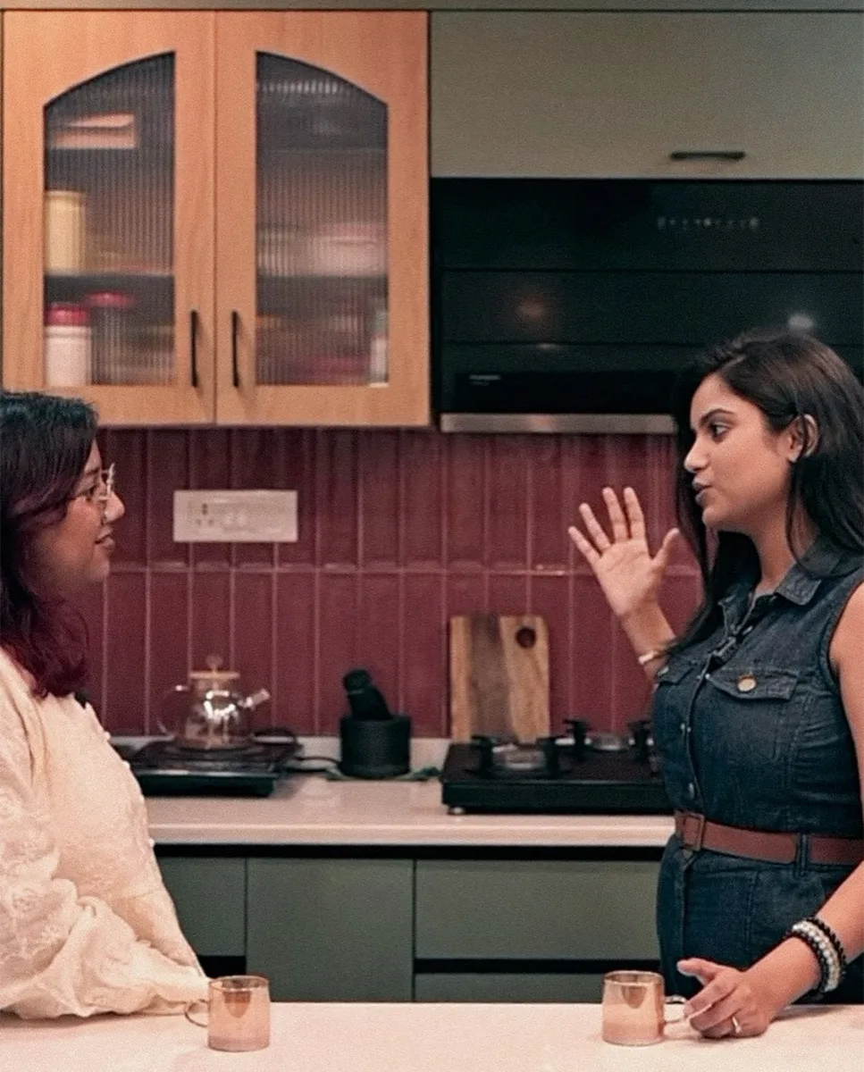 Two women talking across a kitchen counter, sage cabinets and oxblood tiling behind them
